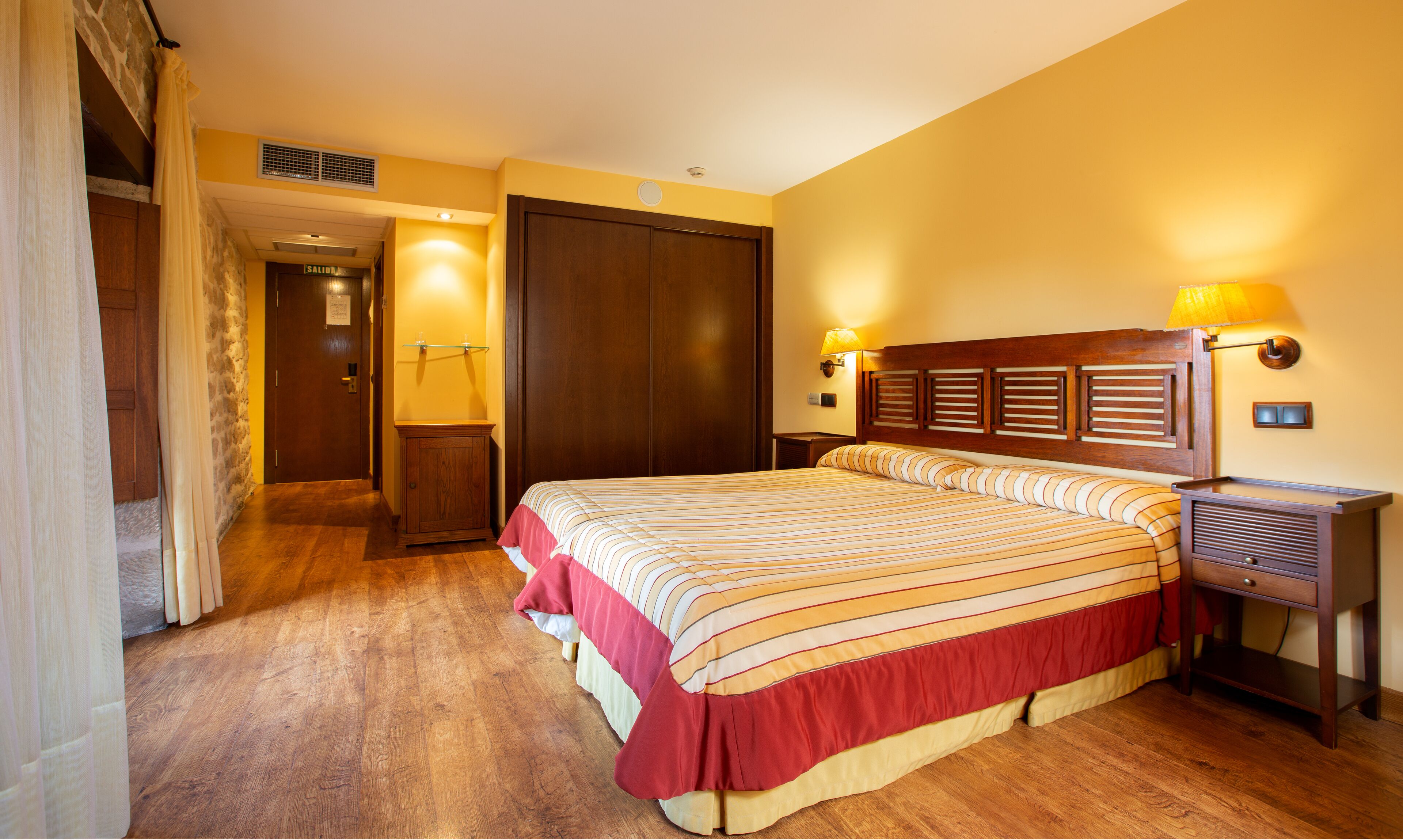 Une chambre d’hôtel avec un grand lit, une tête de lit en bois, deux lampes de chevet, une table de chevet et une armoire.