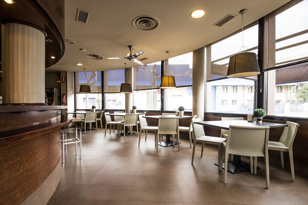 Un interior de restaurante moderno con una barra de madera curvada, varias mesas y sillas, iluminación colgante y grandes ventanales que ofrecen vistas al exterior.