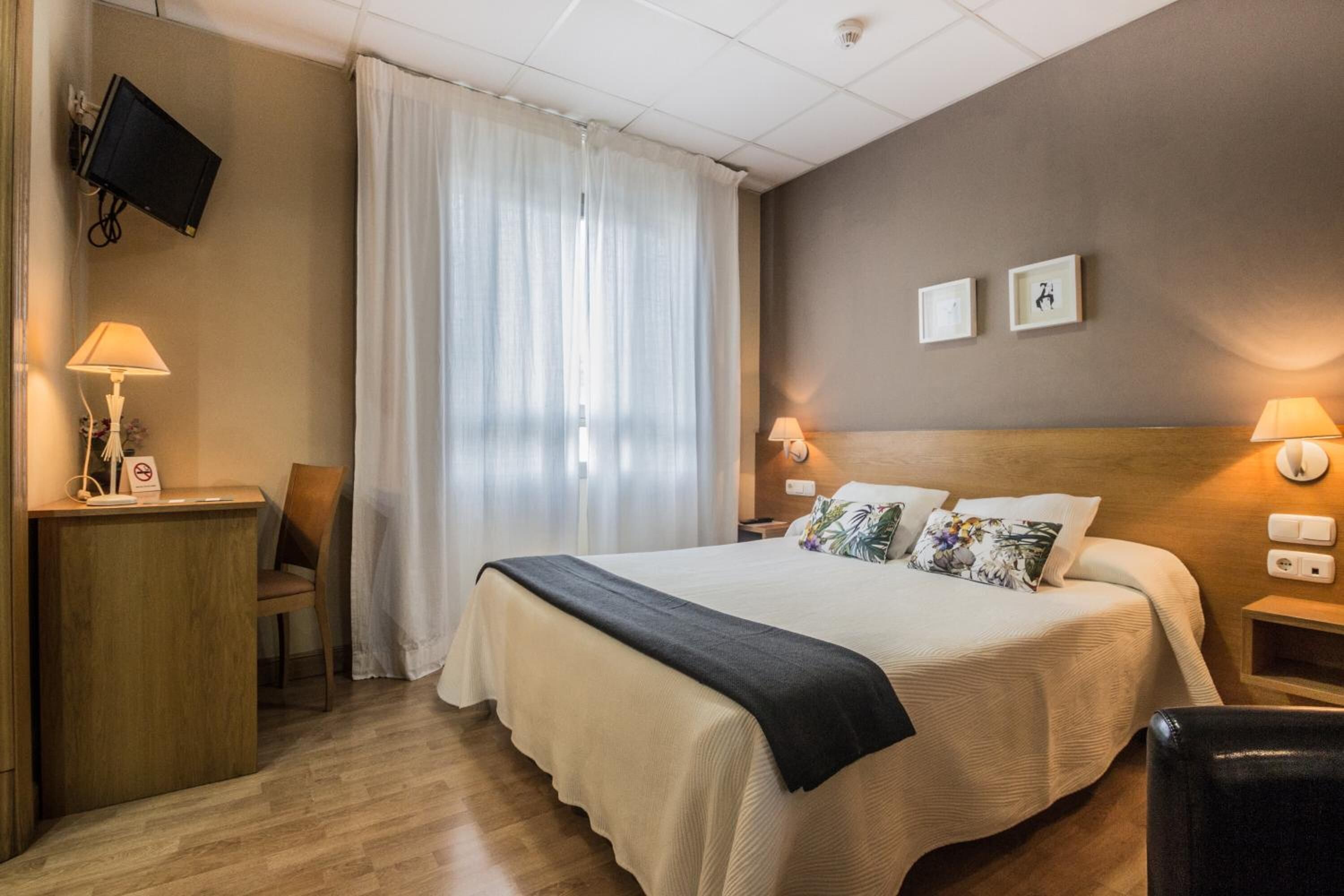 Habitación de hotel con cama, escritorio, silla, televisión y ventana con cortinas.