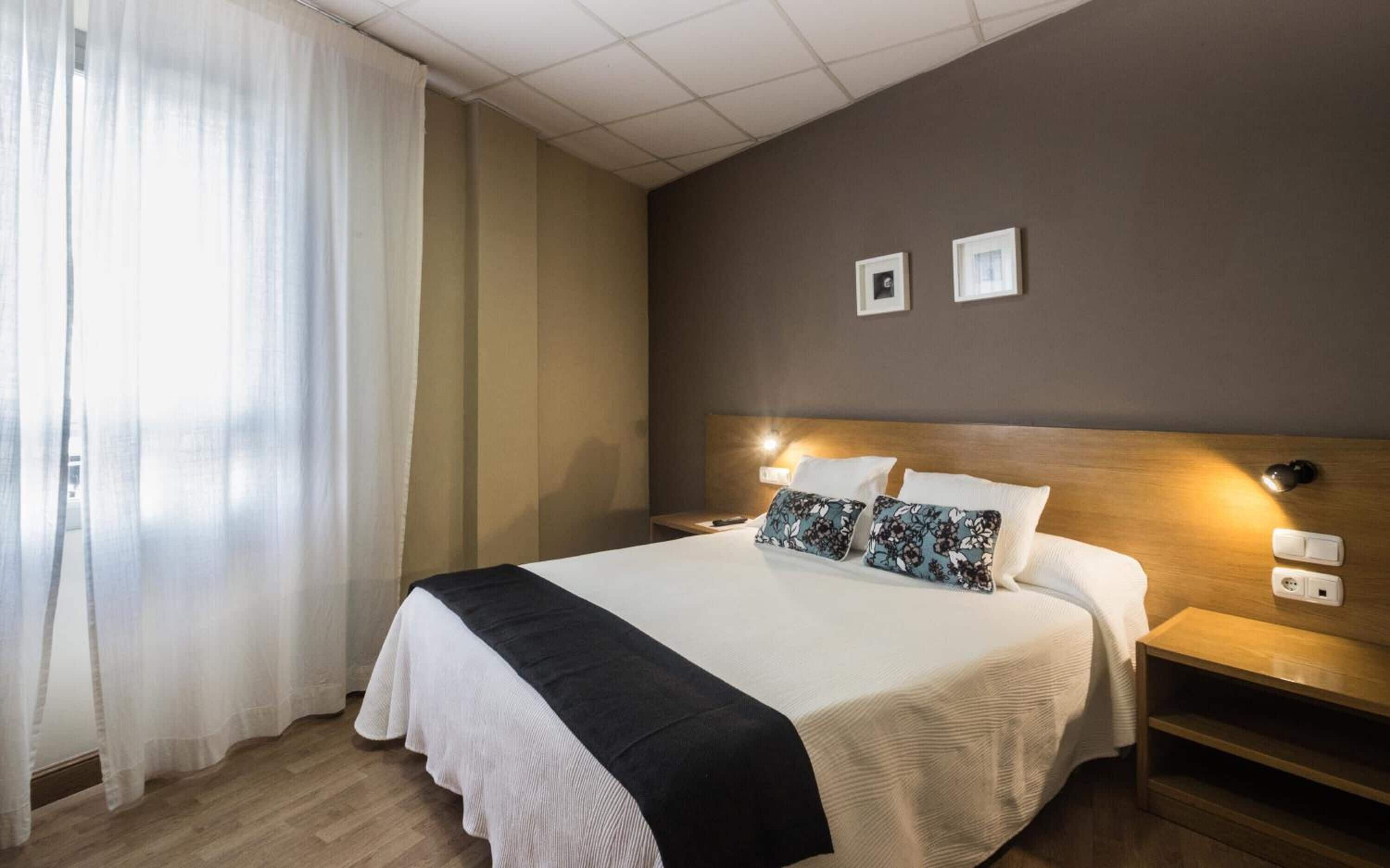 A hotel room with a large bed, two bedside tables, a nightstand, a wooden headboard, and a wall-mounted lamp.