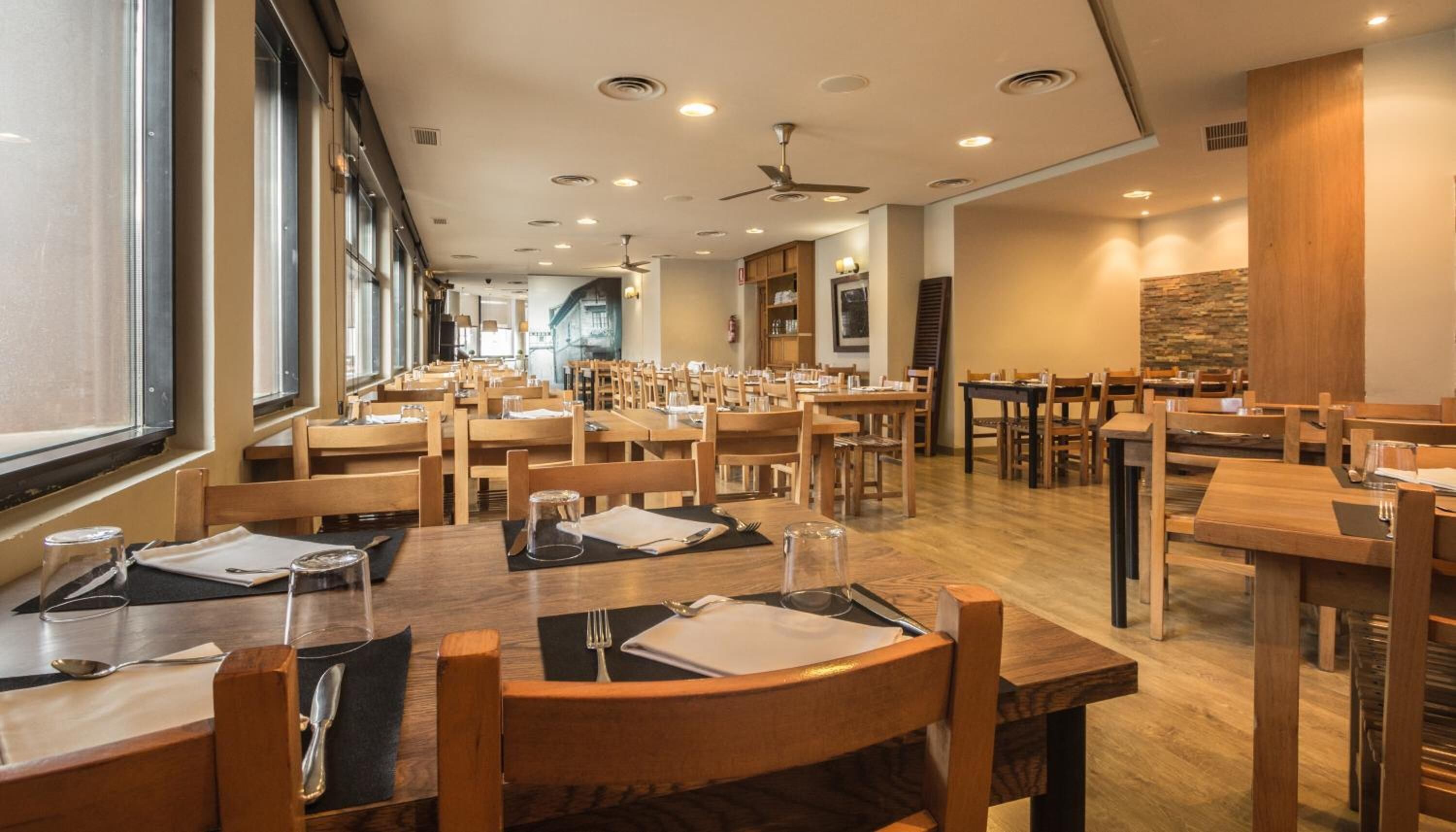 Un restaurante espacioso con mesas y sillas de madera, ventanas amplias y un diseño de techo moderno.