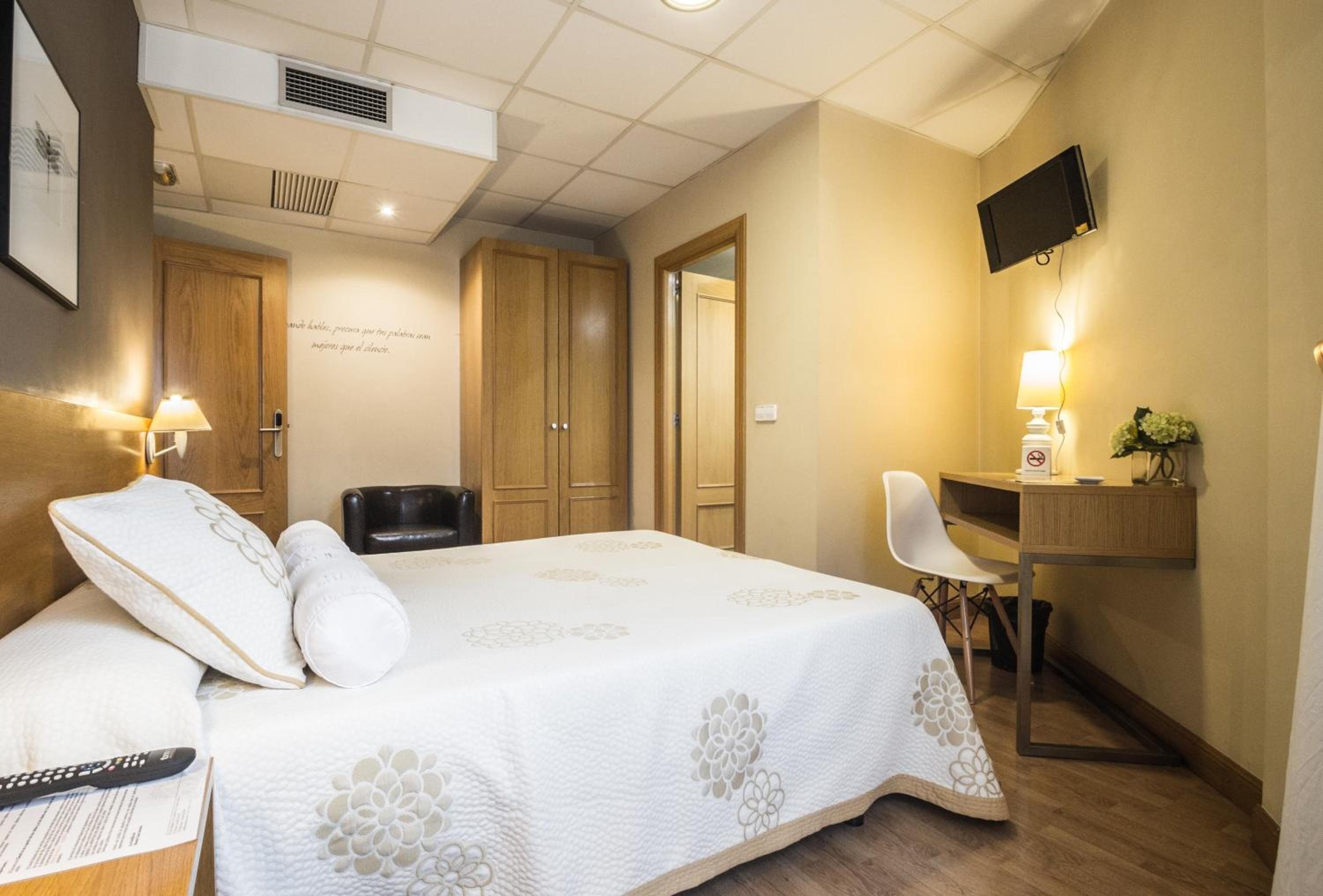 A hotel room with a bed, bedside table, lamp, desk, chair, TV, and wardrobe.