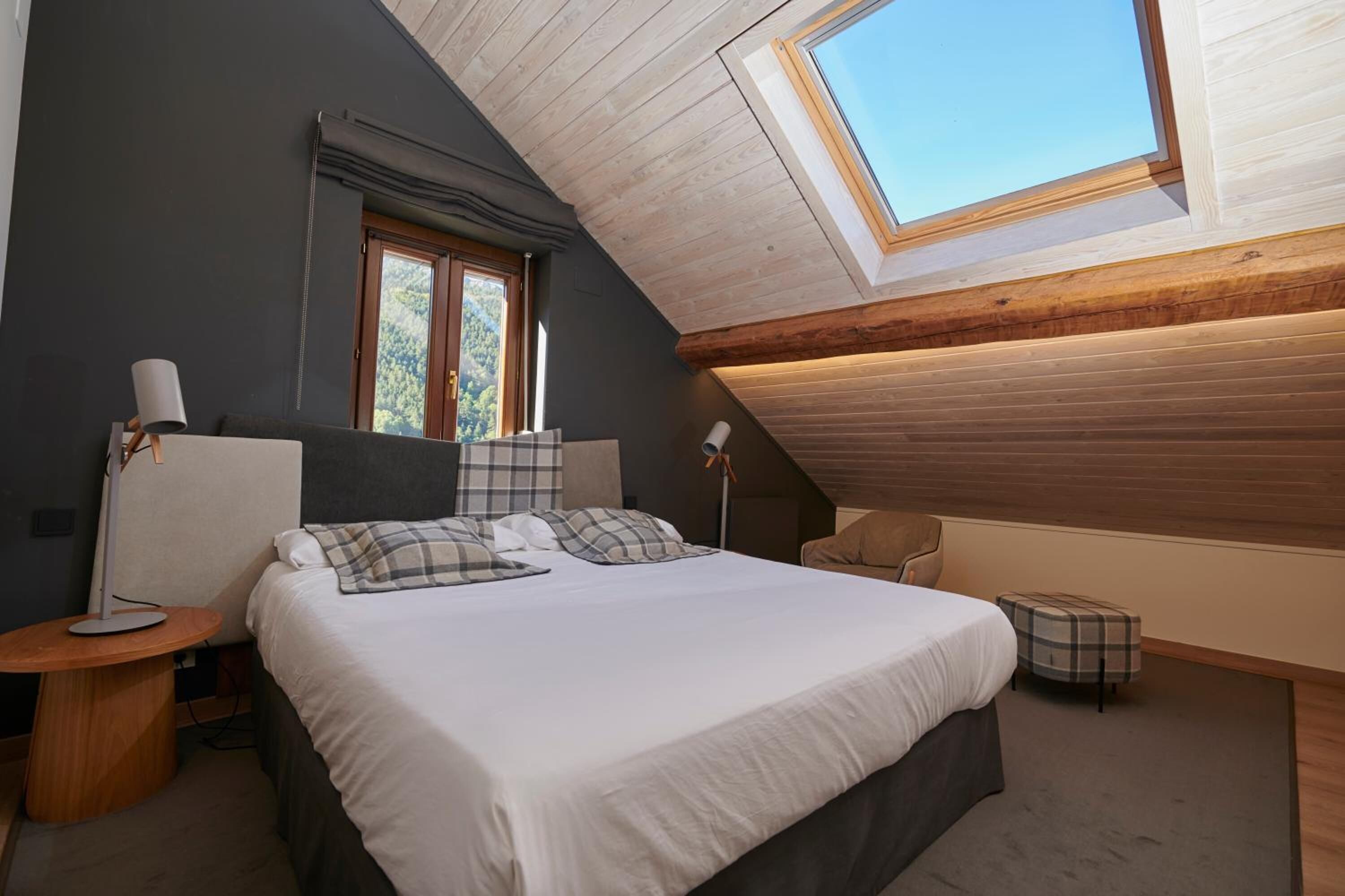 Un dormitorio con tragaluz, una cama con ropa de cama escocesa, una mesita de noche con lámpara y un pequeño otoman.