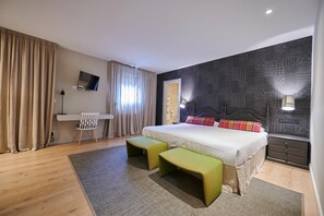 A modern hotel room with a large bed, two green ottomans, a desk with a chair, a flat-screen TV mounted on the wall, and a patterned wall.