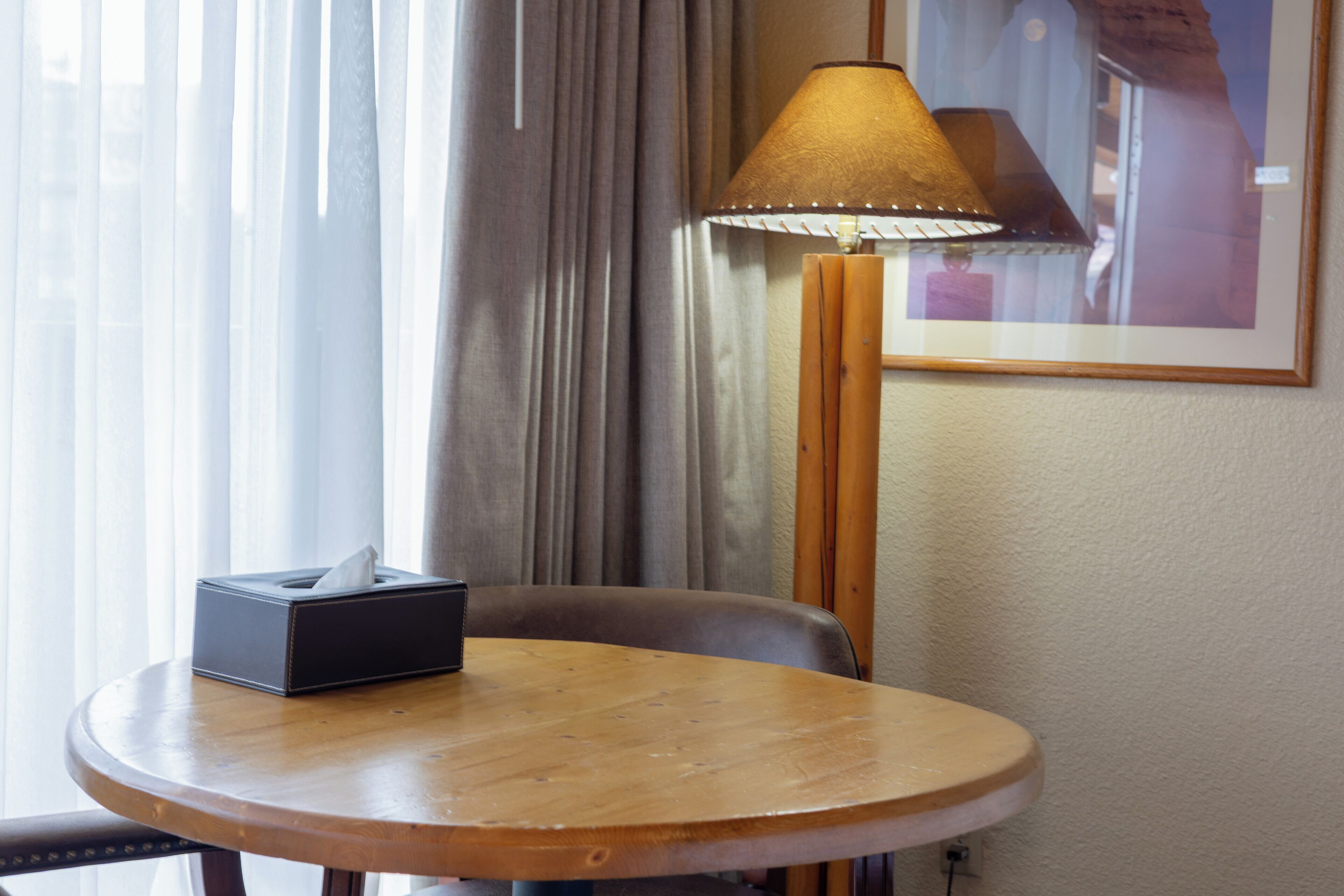 Habitación de hotel con una mesa redonda de madera, una caja de pañuelos, una lámpara y un cuadro en la pared.
