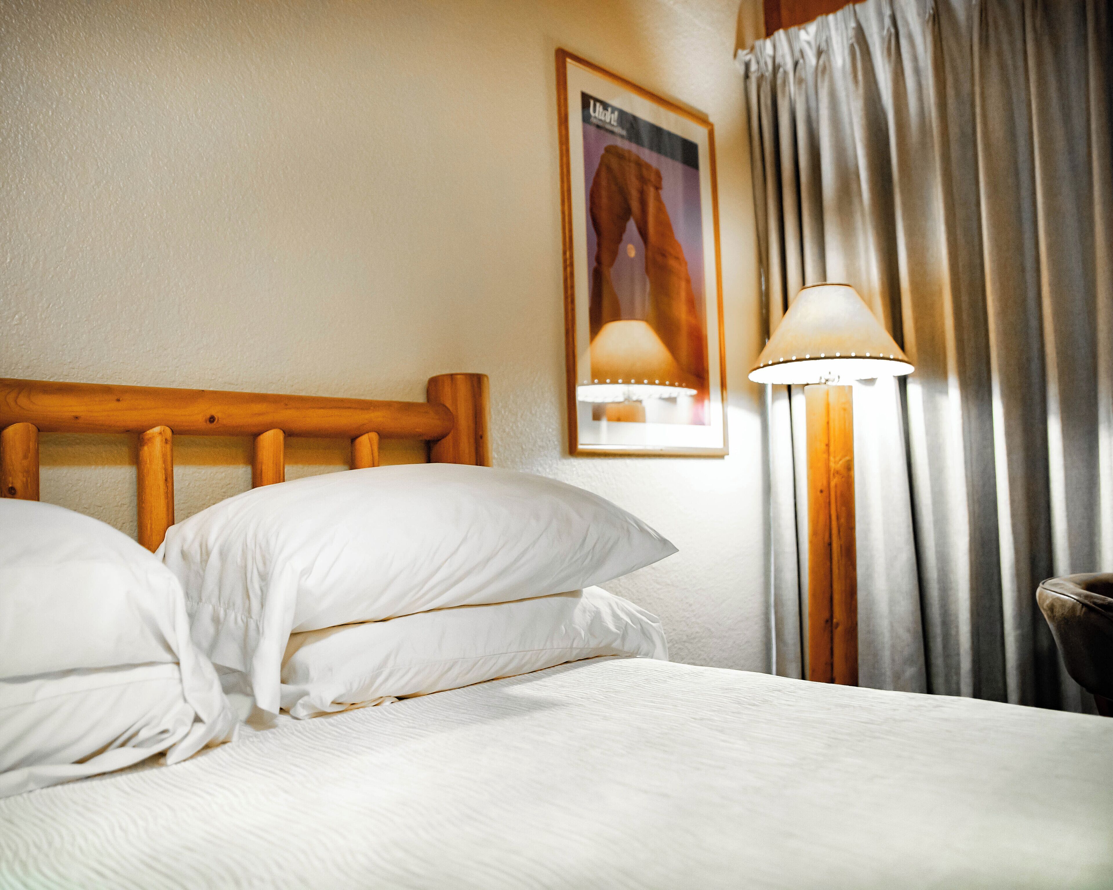 A neatly made bed with white linens, a wooden headboard, and a wall-mounted lamp.