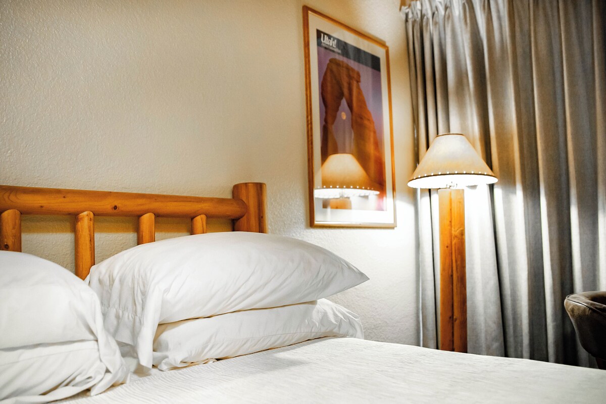 A neatly made bed with white linens, a wooden headboard, and a wall-mounted lamp.