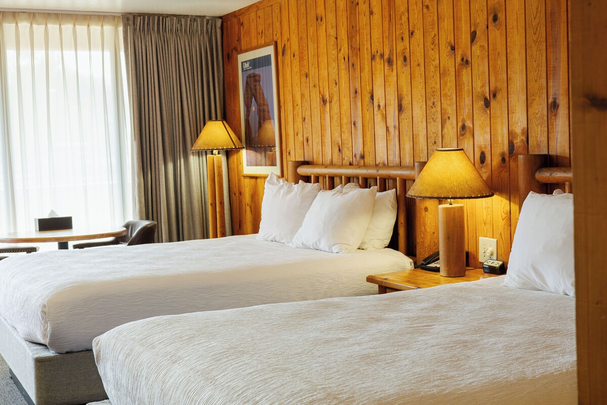A hotel room with two beds, a wooden headboard, a table, and a lamp.