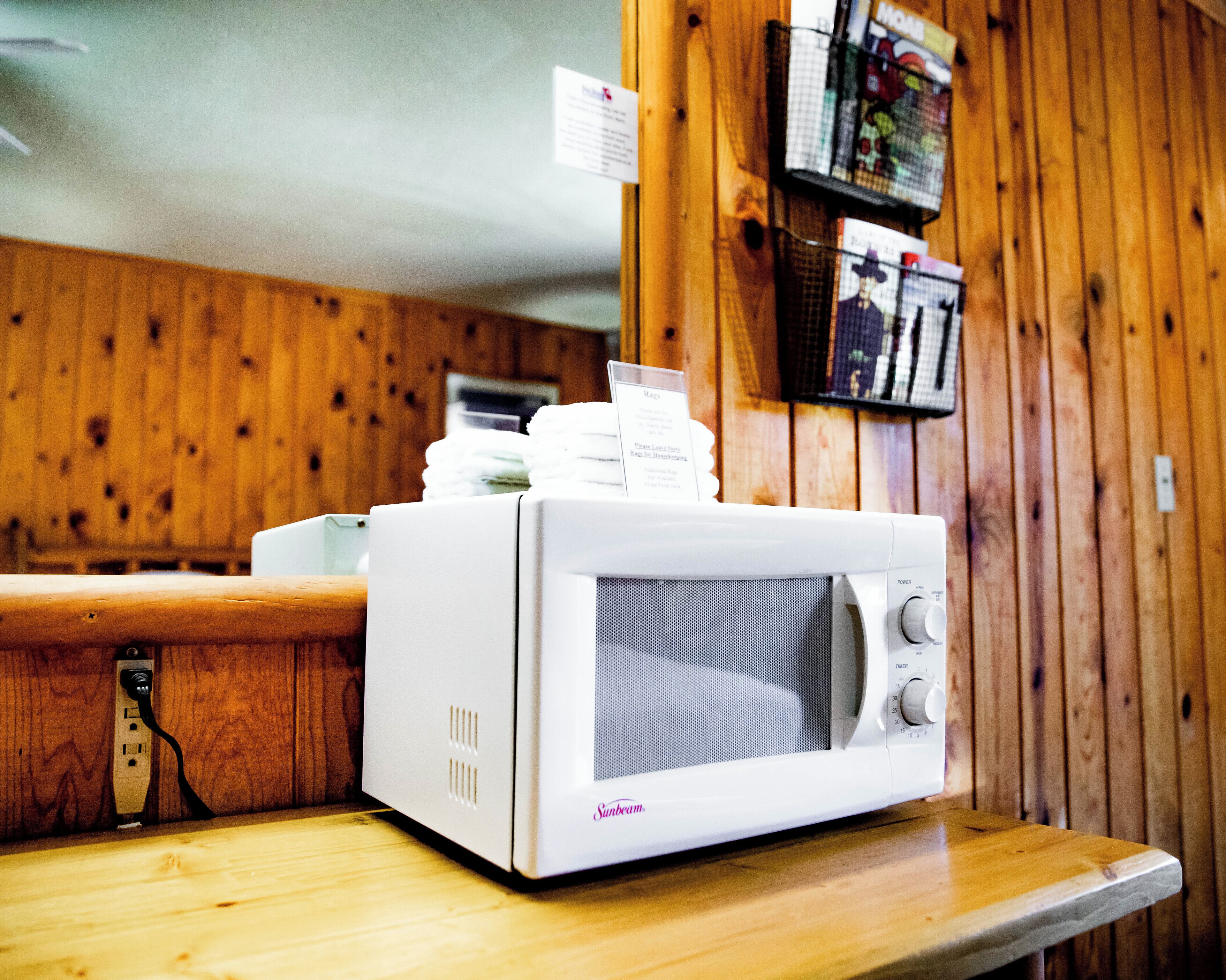 Um micro-ondas branco sobre uma bancada de madeira com uma tomada, uma cesta com revistas fixada na parede e uma parede de madeira com ripas verticais.