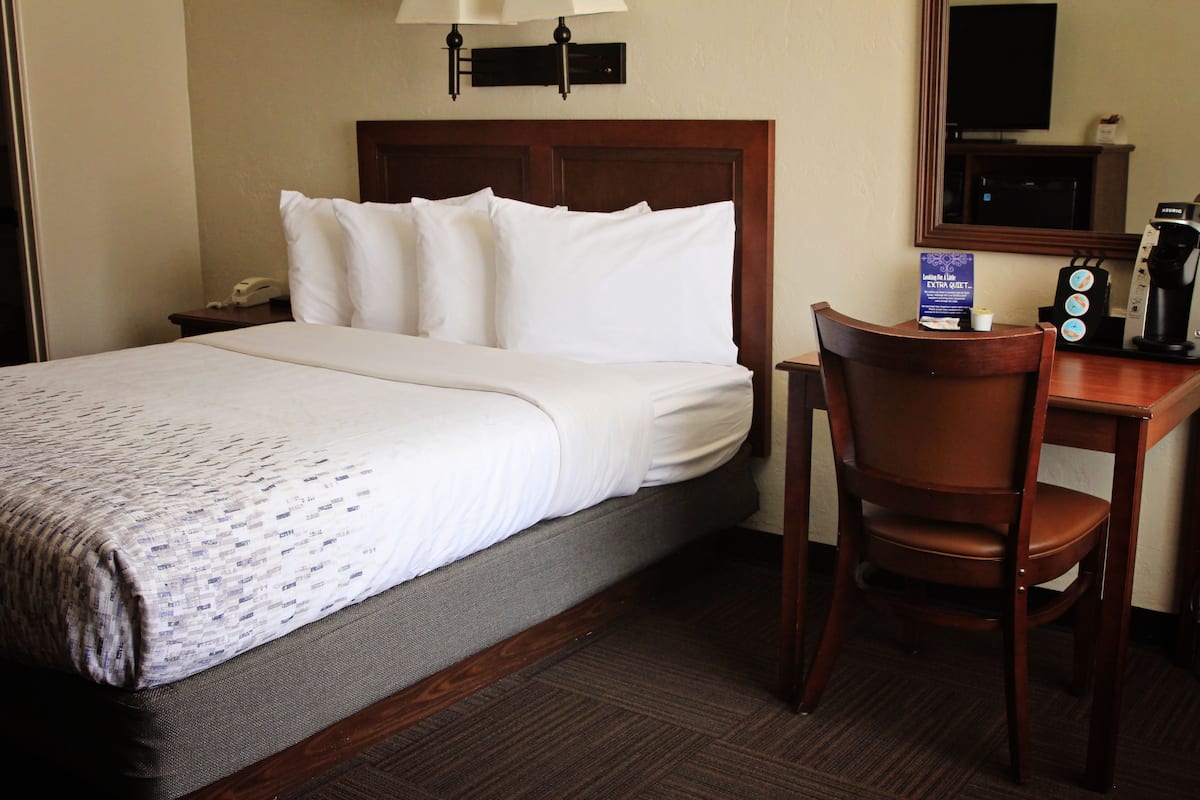 A hotel room with a bed, a desk with a coffee maker, a chair, and a nightstand with a phone.