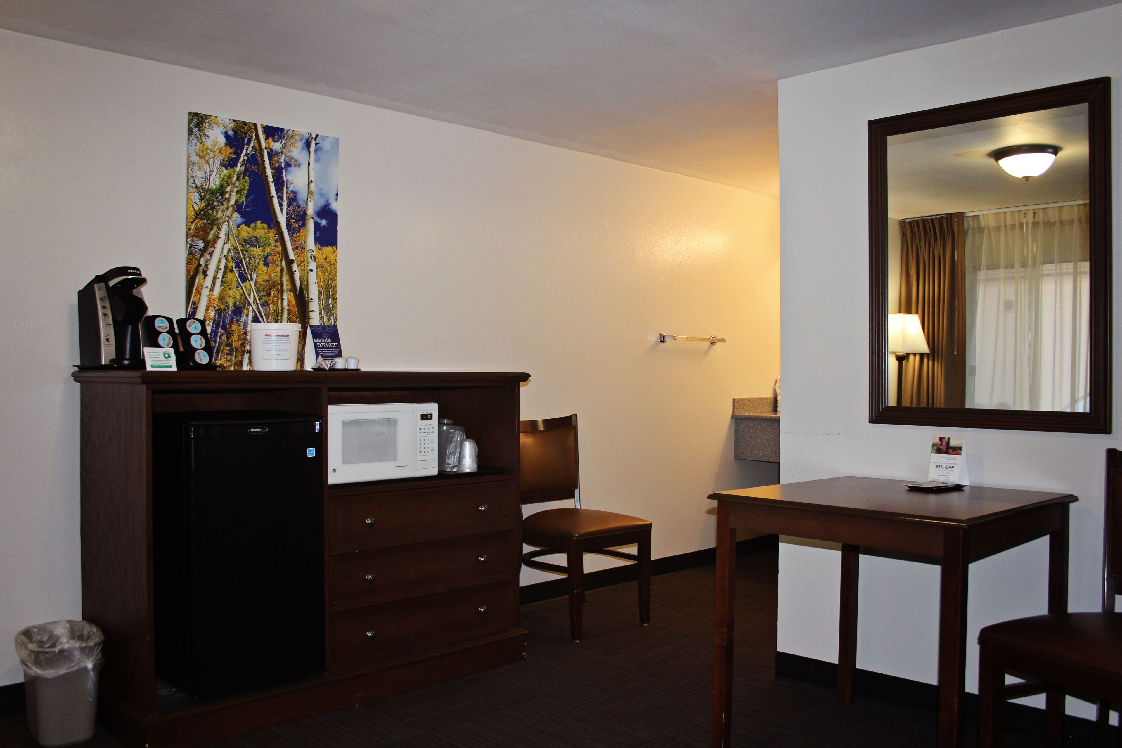 A hotel room with a kitchenette, a microwave, a coffee maker, a trash can, a chair, a table, a mirror, and a painting on the wall.