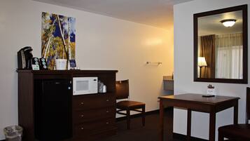 A hotel room with a kitchenette, a microwave, a coffee maker, a trash can, a chair, a table, a mirror, and a painting on the wall.
