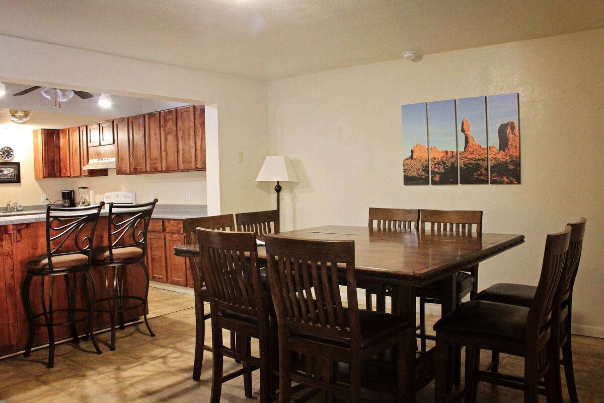 A dining area with a wooden table and chairs, a kitchen in the background, and a desert landscape painting on the wall.