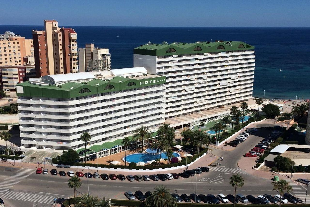 Et hotel med flere etager, en pool på taget og palmer, omgivet af en parkeringsplads.