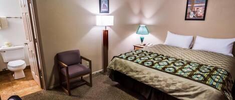 A hotel room with a bed, a chair, a bedside table with a lamp, a toilet, and a picture on the wall.