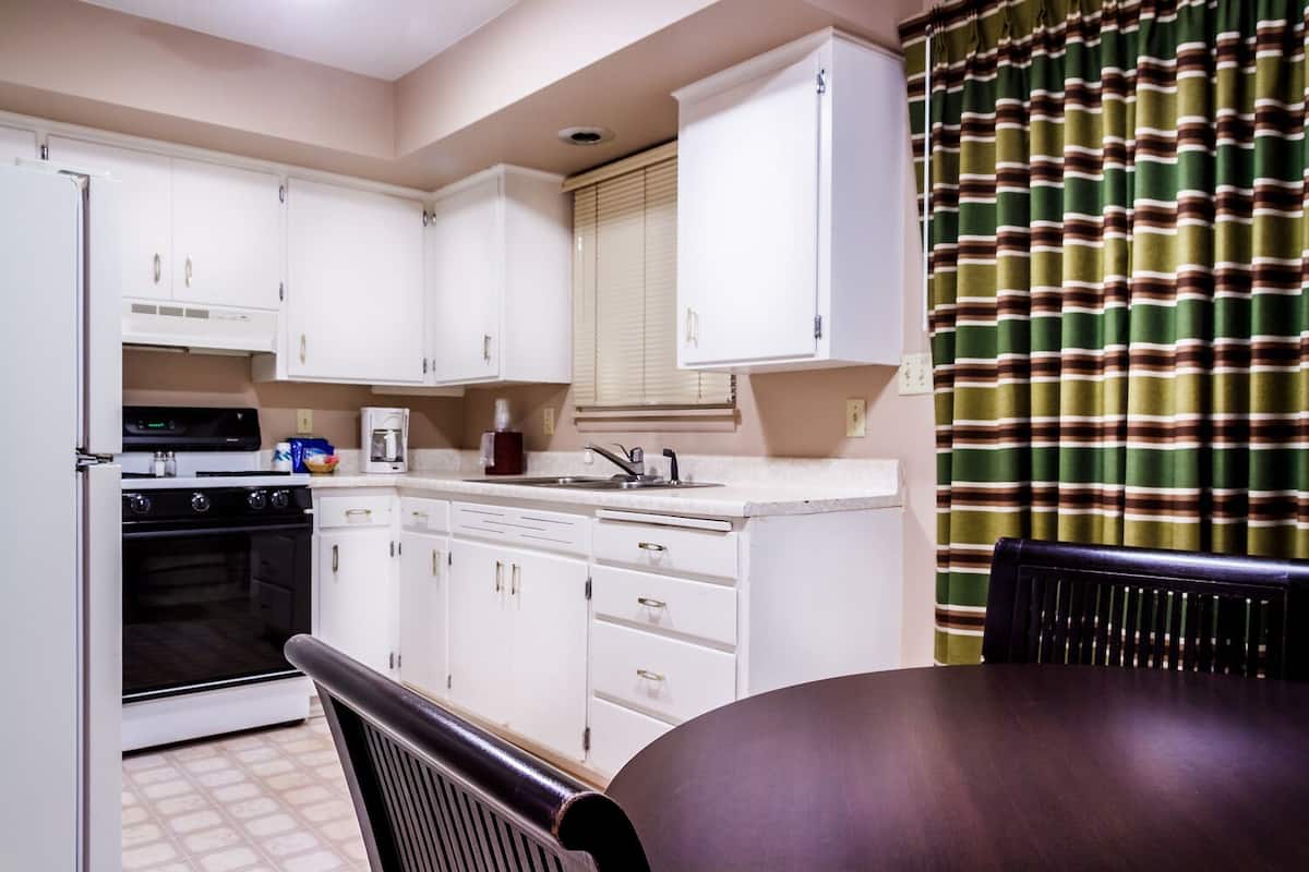 Une cuisine compacte avec des meubles blancs, une cuisinière et un lave-vaisselle. On y trouve une table à manger avec une chaise.