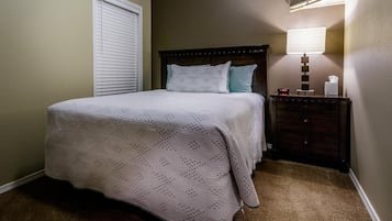Un lit bien fait, recouvert d’une couverture blanche, avec une tête de lit en bois foncé et une table de chevet sur laquelle se trouvent une lampe et une horloge.
