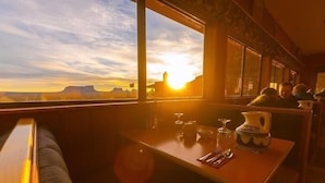 Un wagon-restaurant avec une table dressée pour deux personnes, une fenêtre donnant sur un paysage où le soleil se couche derrière une chaîne de montagnes, et une personne assise à la table.