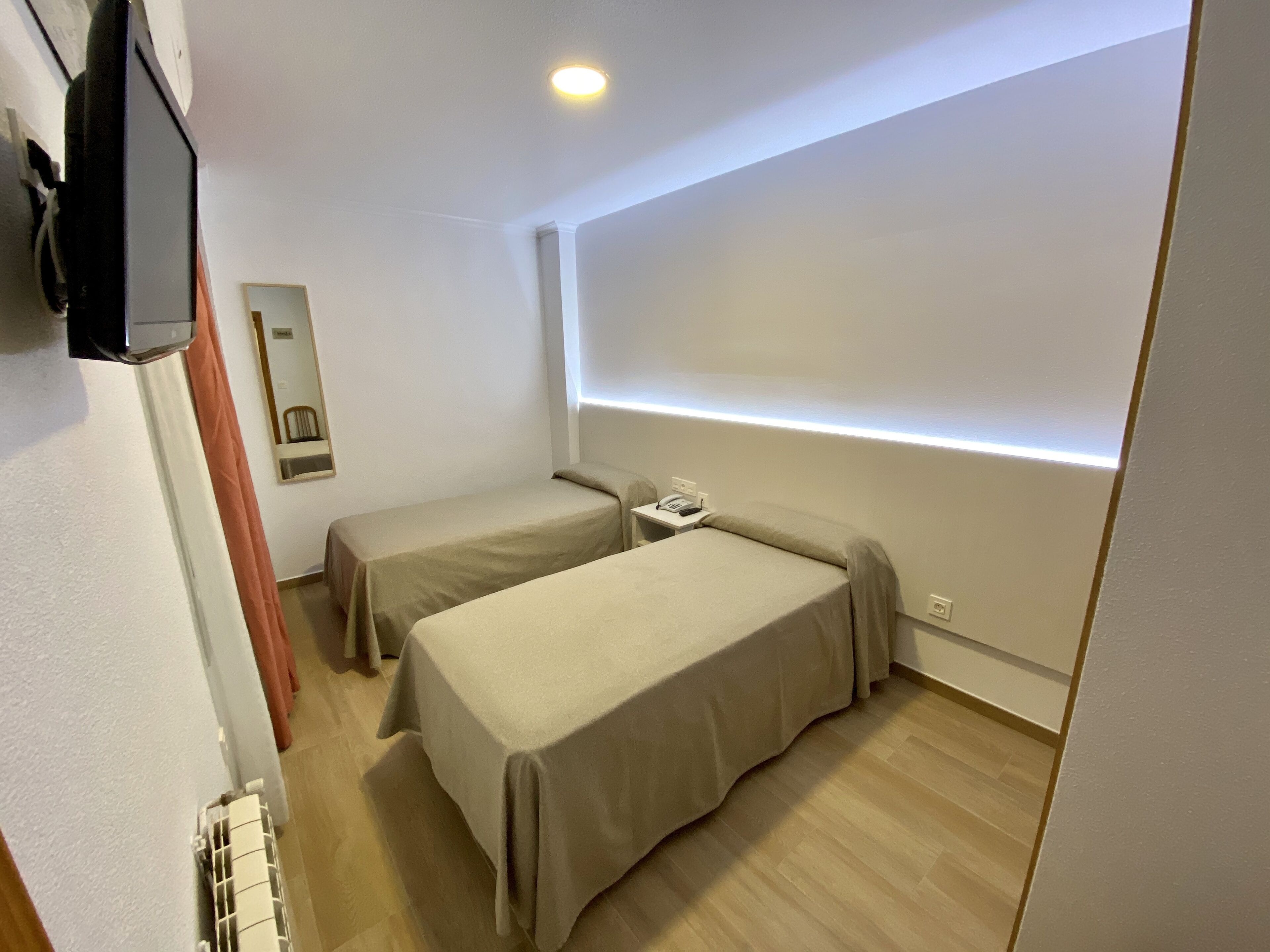 Une petite chambre moderne, équipée de deux lits simples, d’une table de chevet avec un téléphone et d’un téléviseur à écran plat fixé au mur.