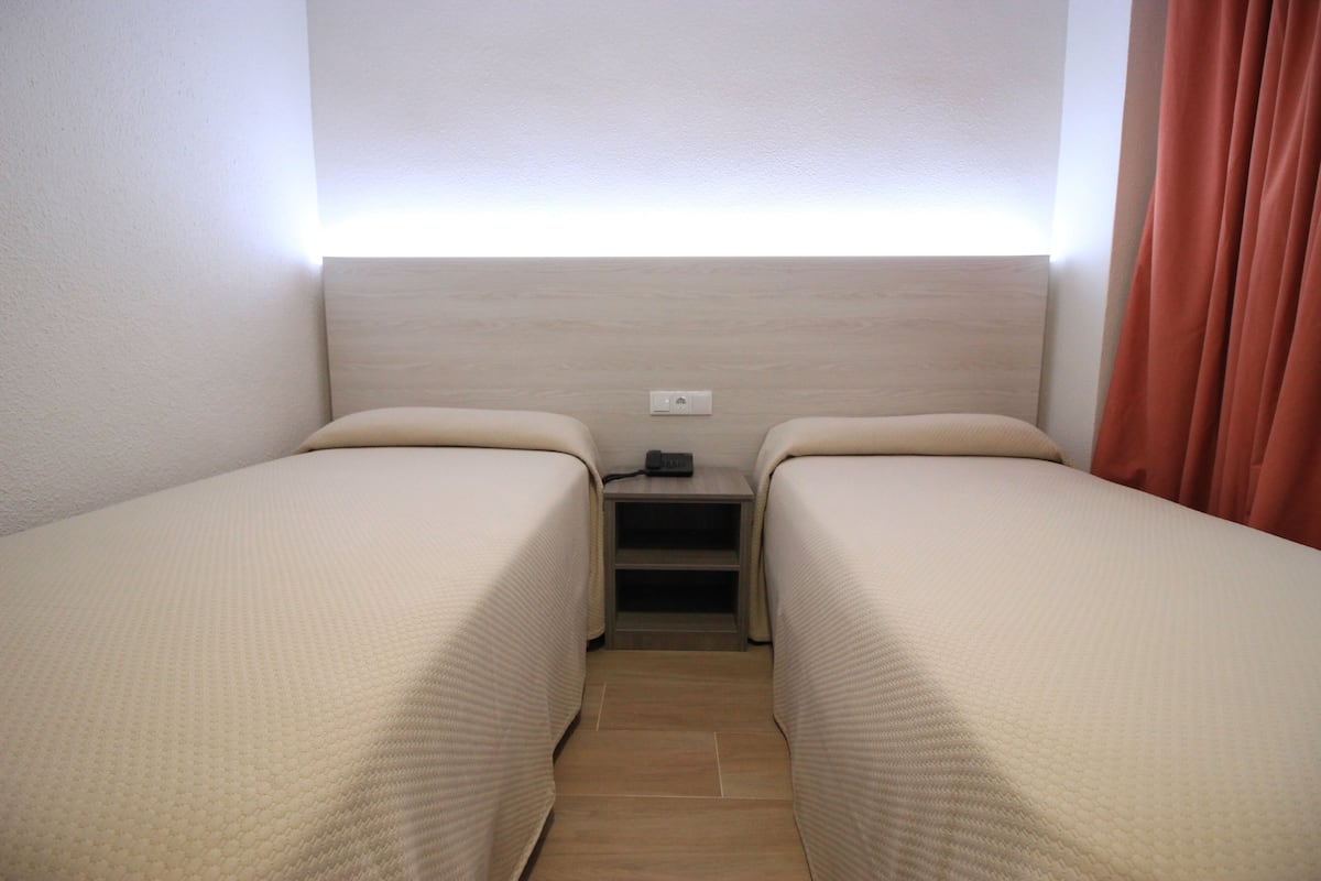 A small, well-lit room with two single beds, a nightstand, and a power outlet.
