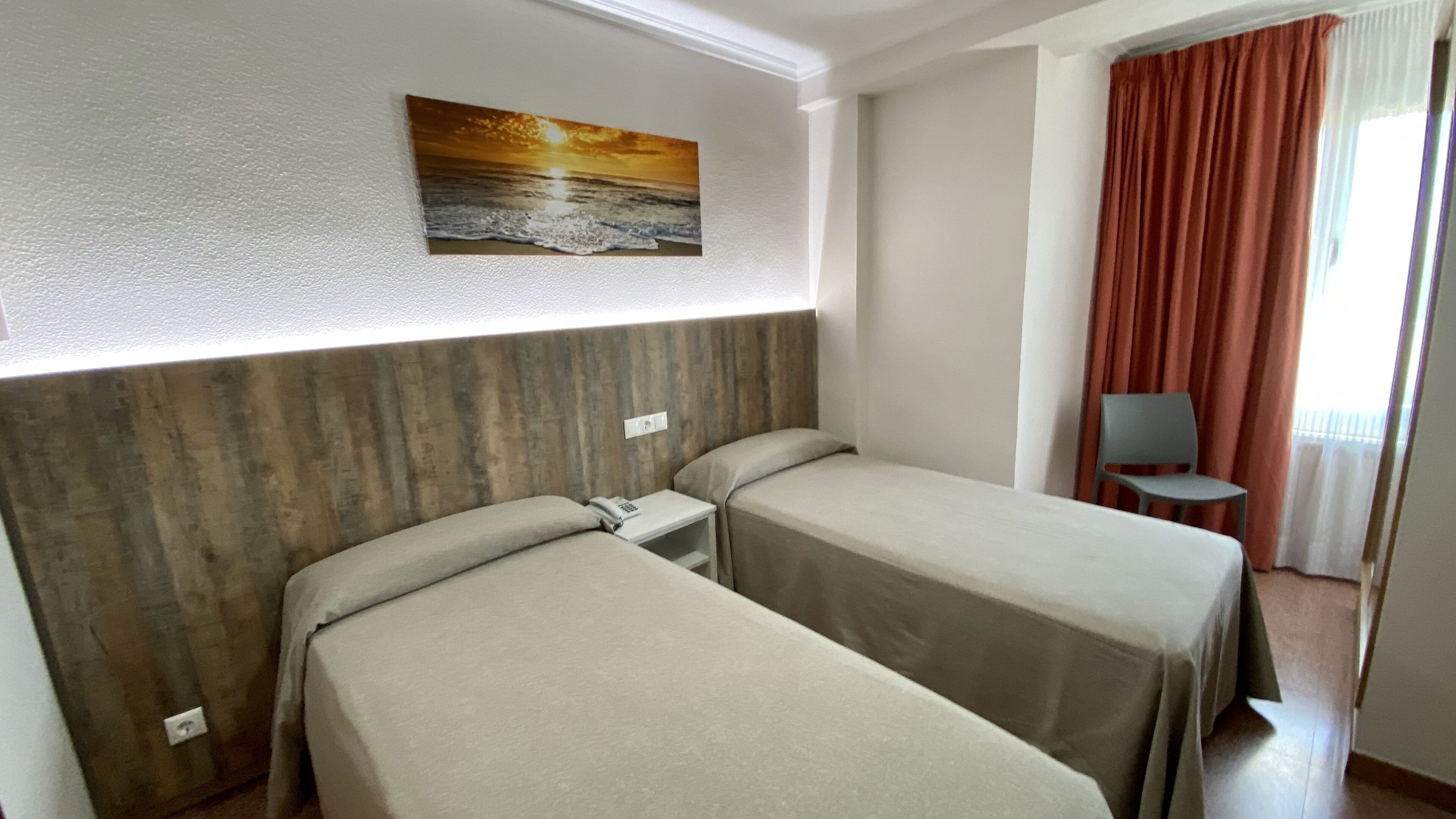 Ein Hotelzimmer mit zwei Einzelbetten, einem hölzernen Kopfteil, einem Sonnenuntergangsgemälde, einem Schreibtisch mit Telefon und einem Stuhl an einem fensterbelzogenen Fenster.
