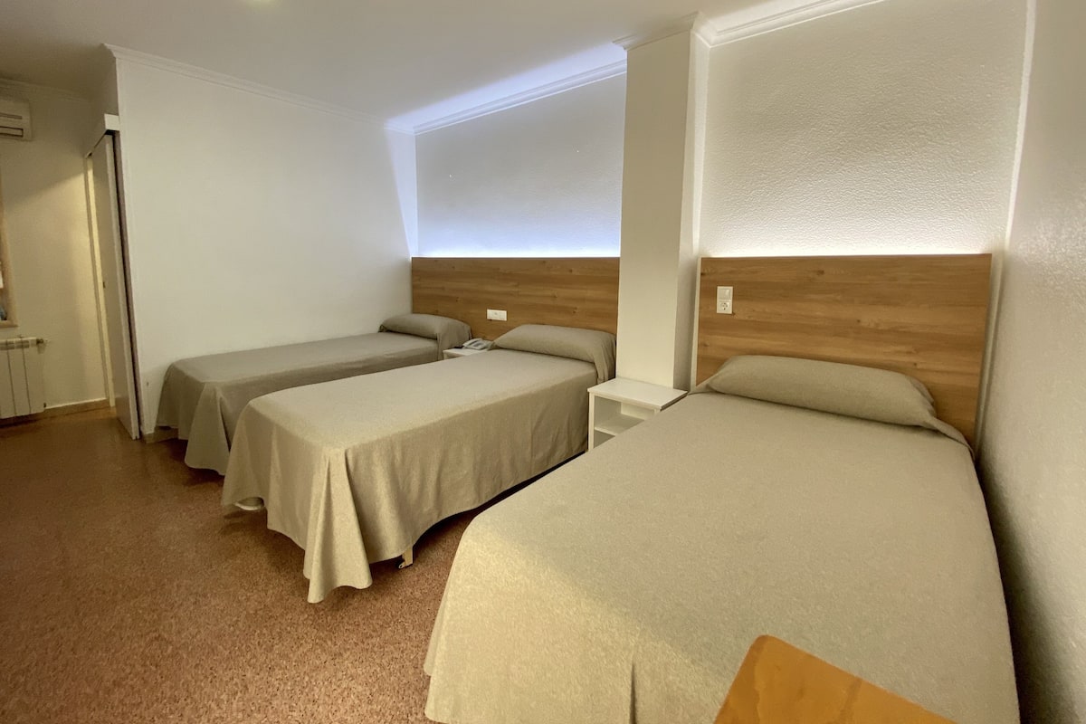 A hotel room with three single beds, a small bedside table, and a window with a view.