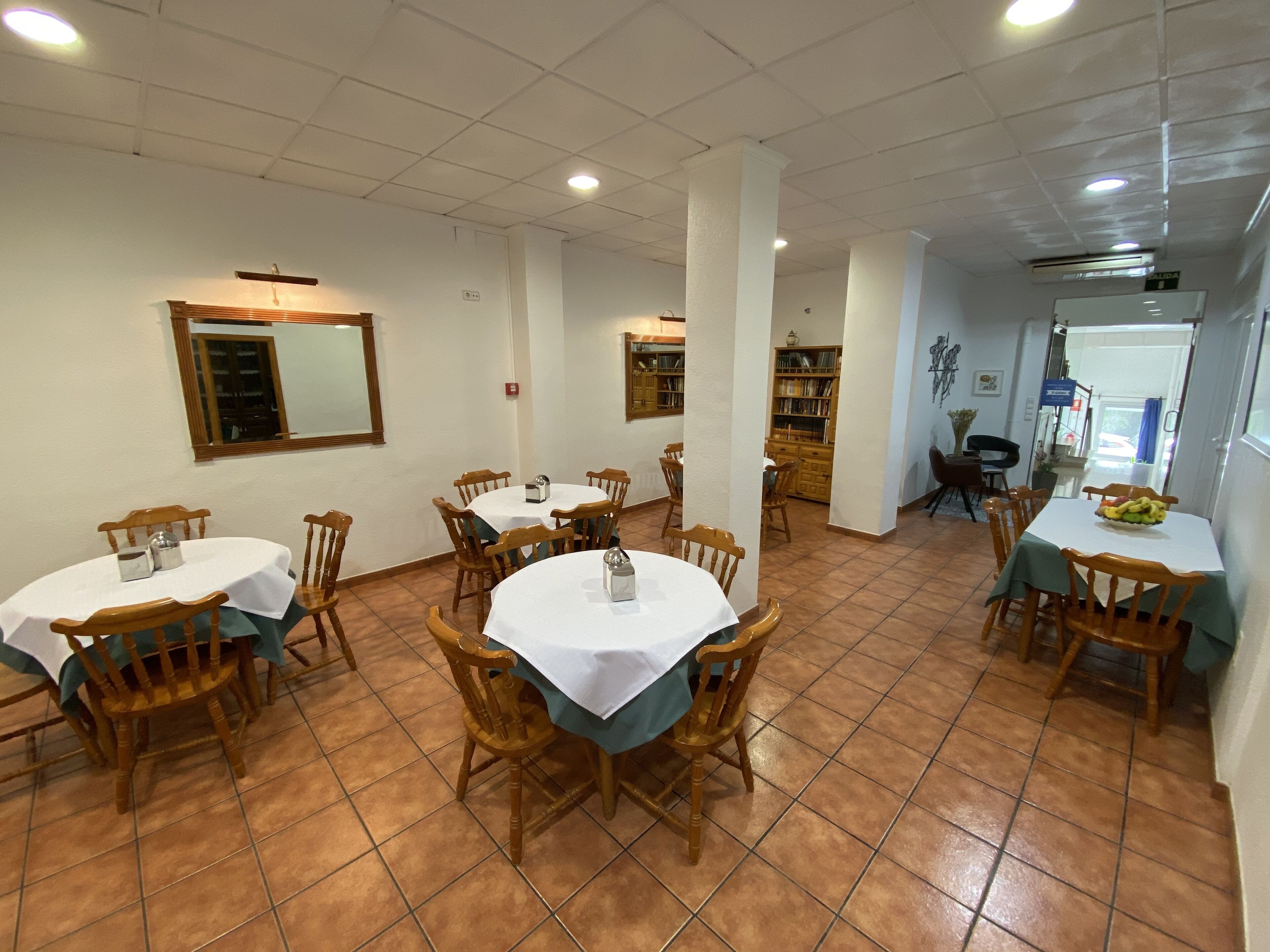Un comedor con mesas redondas y sillas de madera, suelo de baldosas y un espejo en la pared.