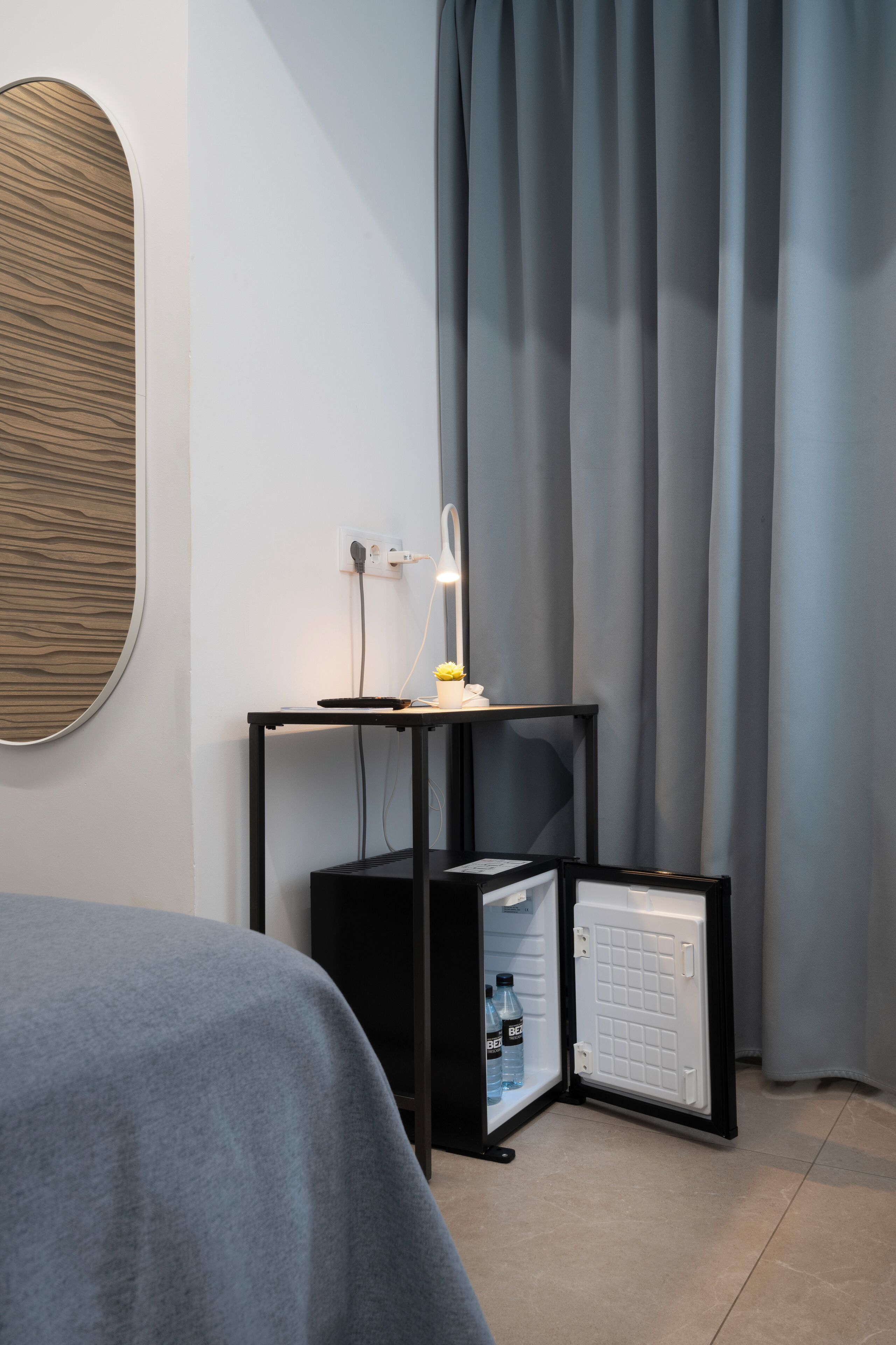 Ett hotellrum med en säng, ett litet bord med en lampa, ett minikylskåp och en spegel som är monterad på väggen.