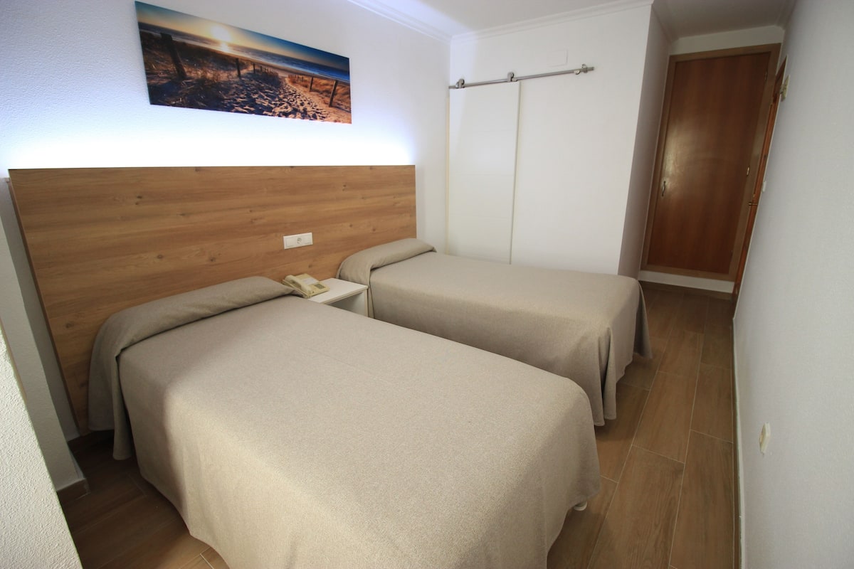 A hotel room with two beds, a wooden headboard, a nightstand, a painting on the wall, and a wooden door.