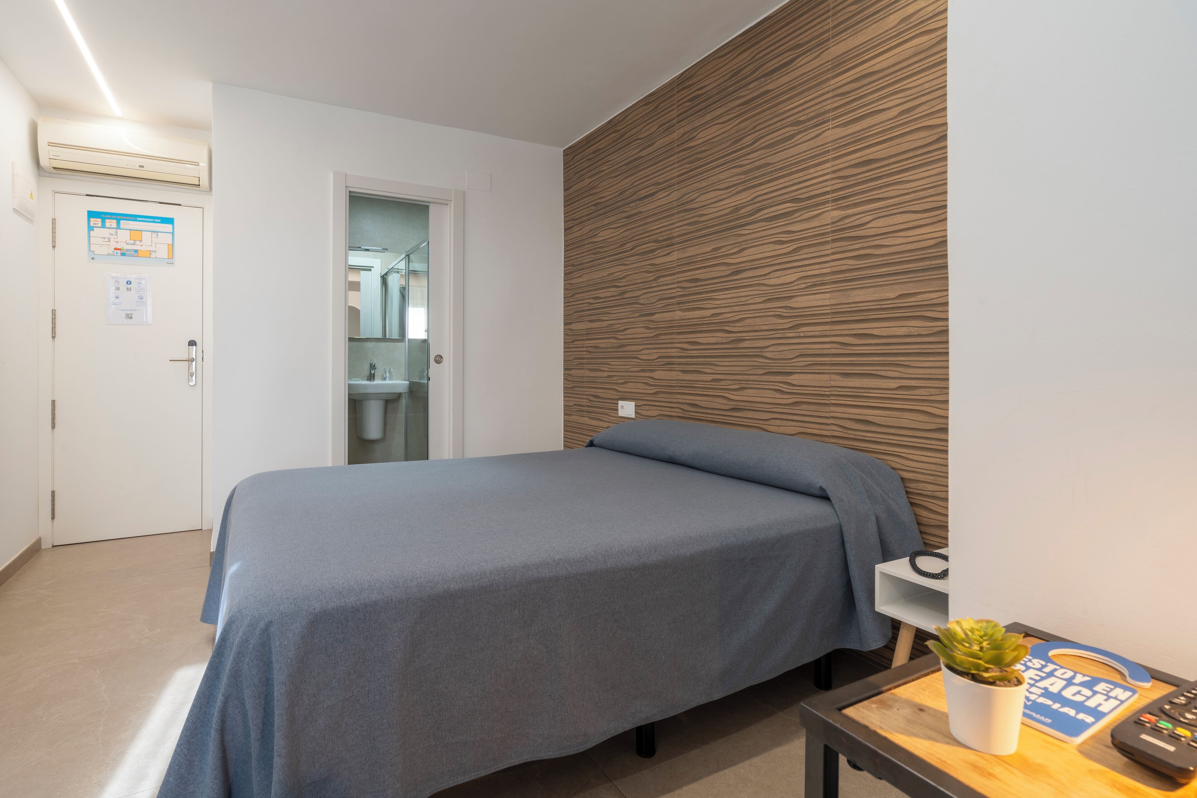 Un dormitorio con cama, un panel decorativo de madera, una mesita con una planta y un control remoto.