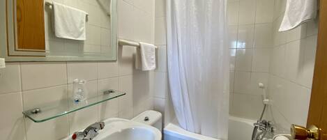 Un baño con lavamanos, inodoro y bañera blancos. Hay un estante de vidrio sobre el lavamanos y un perchero en la pared.