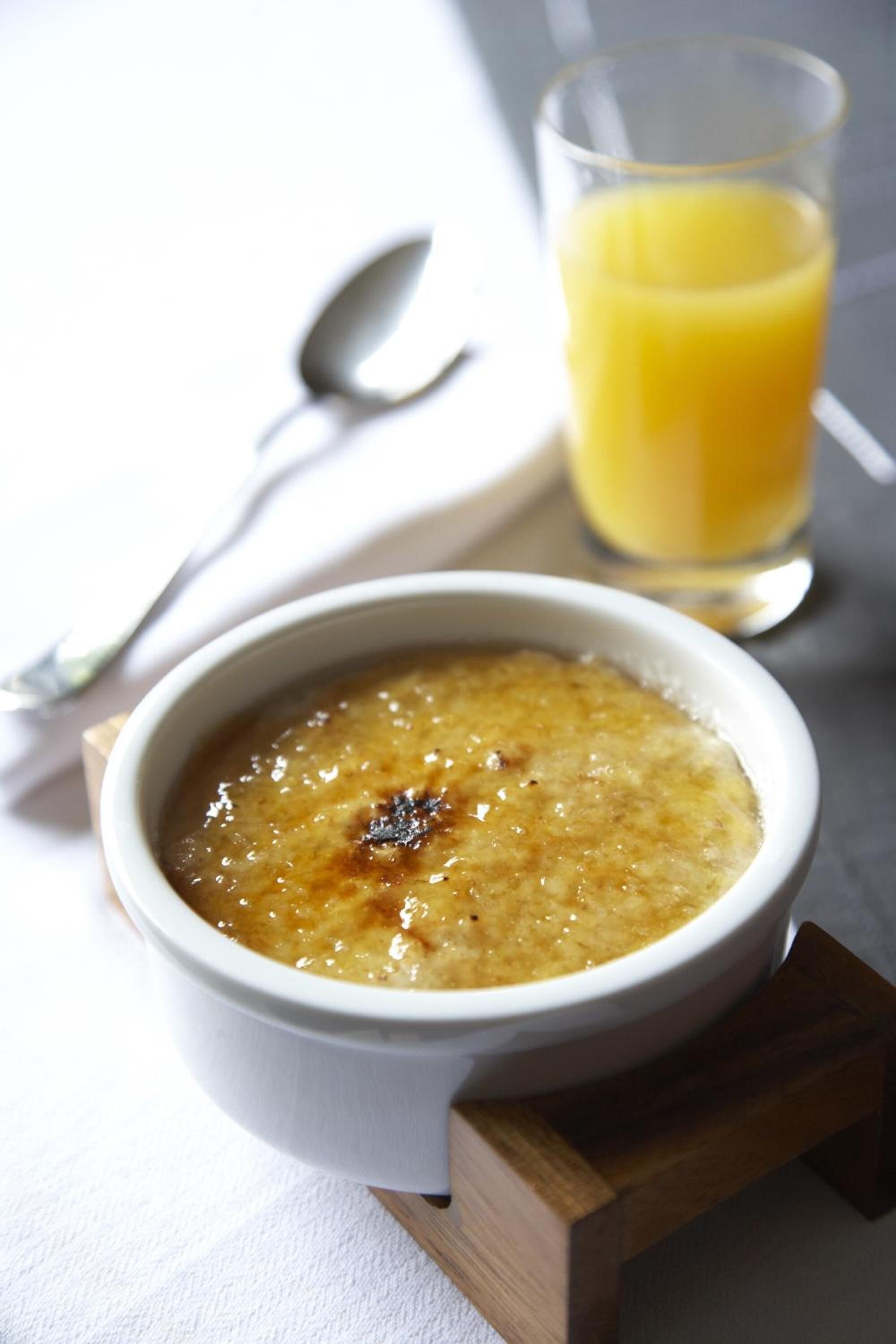 A bowl of crème brûlée on a wooden stand with a glass of orange juice in the background.