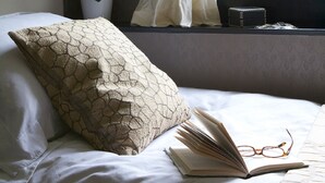 Una cama con un almohadón estampado, un libro abierto y un par de gafas encima.