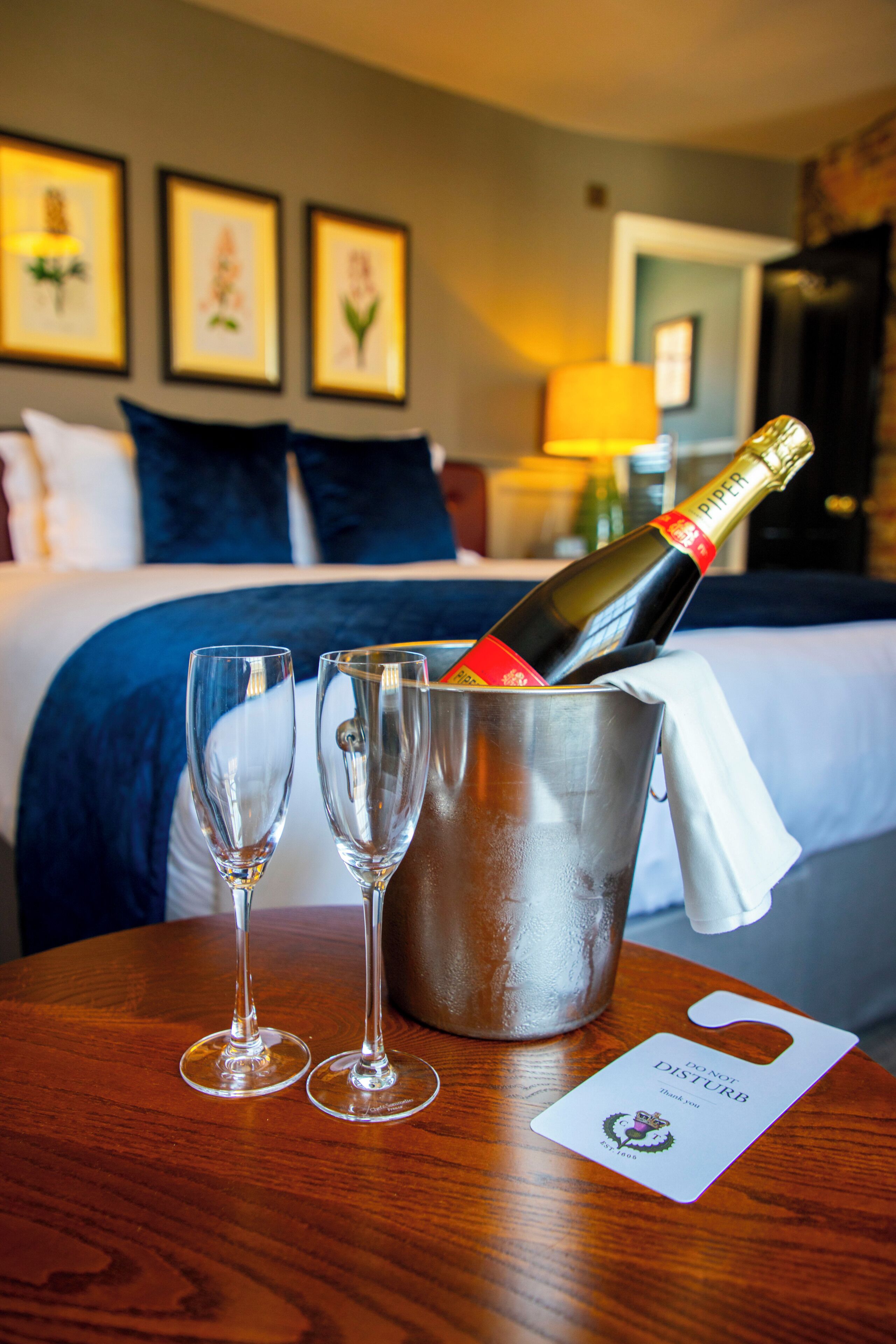 Habitación de hotel con cama, un balde con una botella de champán, dos copas y un cartel de 'no molestar' sobre una mesa de madera.
