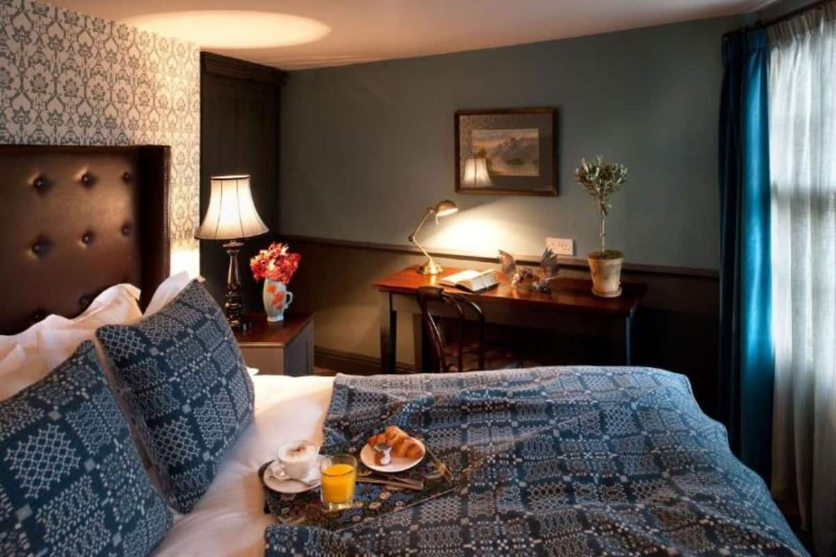 Une chambre d’hôtel avec un lit, un bureau, une chaise, une lampe, un tableau au mur et une fenêtre avec des rideaux.