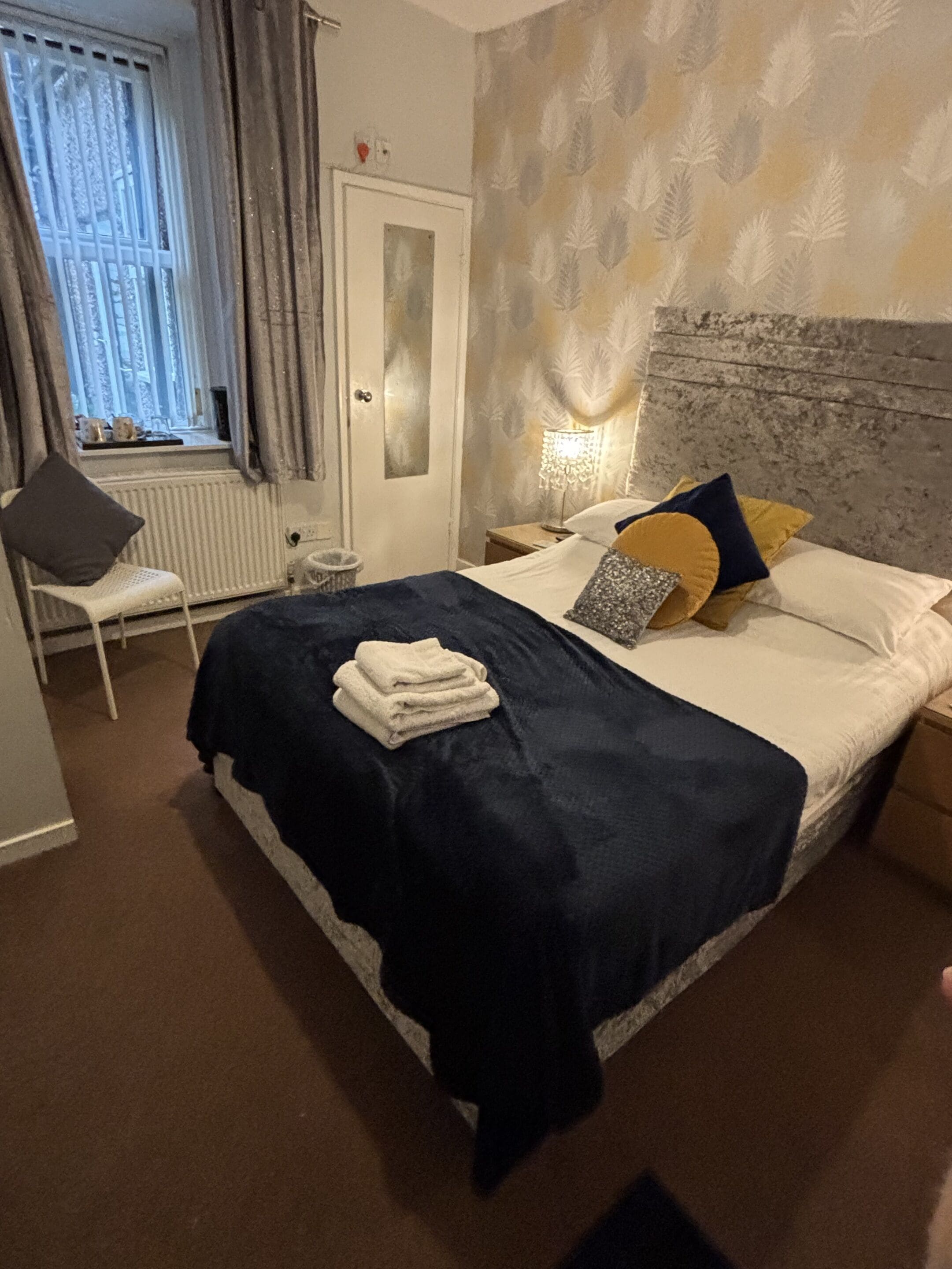A bedroom with a bed, a chair, a nightstand, a lamp, and a window with curtains.