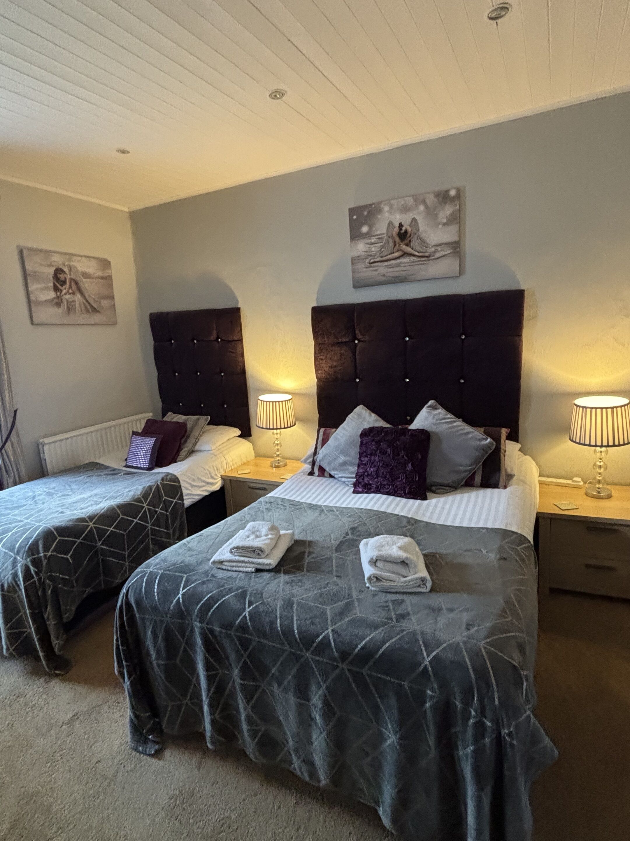 A hotel room with two beds, each with a headboard and bedside lamps. There are towels on the beds and artwork on the walls.