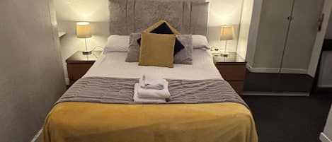 Una cama bien hecha con cabecera, dos mesitas de noche con lámparas, un armario y una obra de arte colgada en la pared.