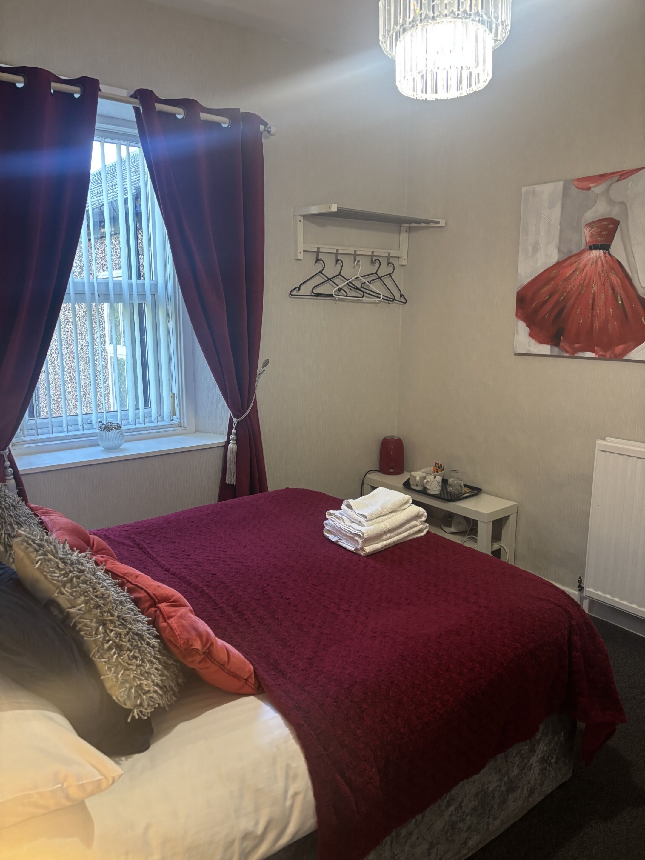 Une chambre avec un lit, une fenêtre avec des rideaux, un radiateur, une petite table avec une bouilloire et un tableau au mur.