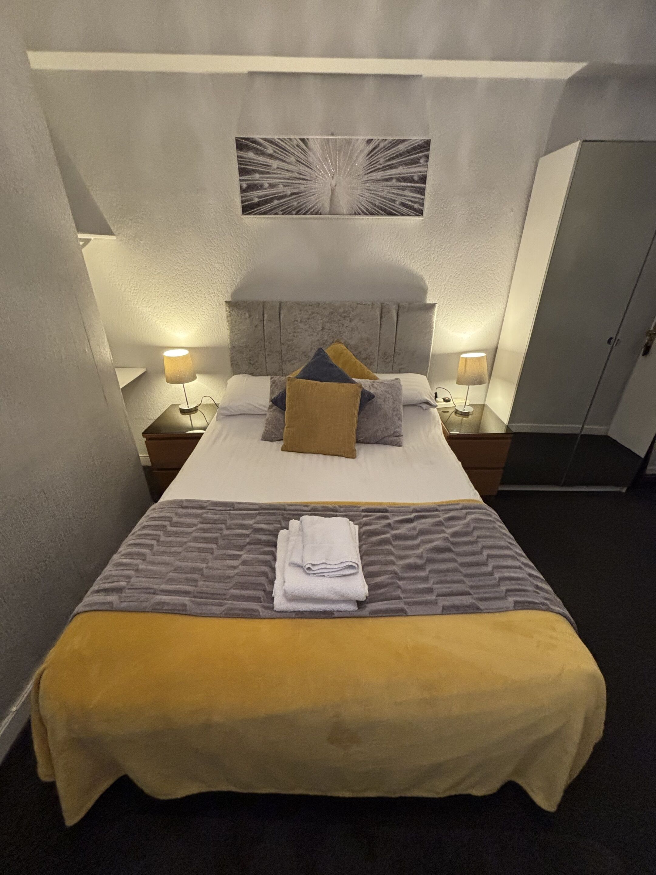 A neatly made bed with a headboard, two bedside tables with lamps, a folded towel on the bed, and a door to the right.