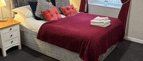 Una cama impecable con cabecera capitonada, una manta roja y una pila de toallas blancas. Se ve una mesita de noche con una lámpara, una ventana con cortinas y vistas a la vegetación.