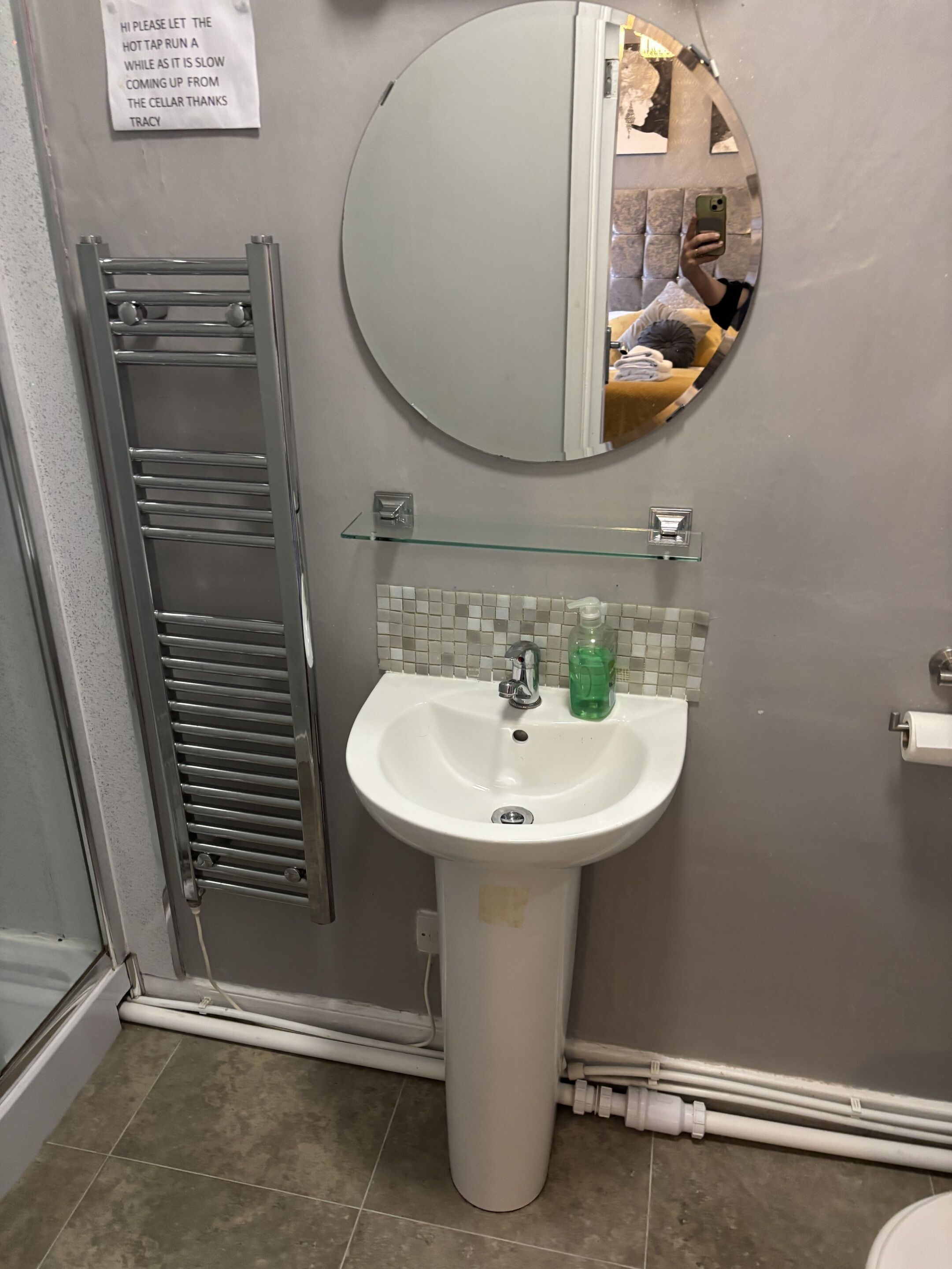 A bathroom with a white pedestal sink, a glass shelf above it, a towel rack, and a mirror with a reflection.