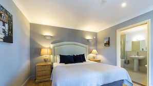 A neatly arranged bedroom with a large bed, two bedside tables with lamps, a wooden dresser, and a bathroom visible through an open door.