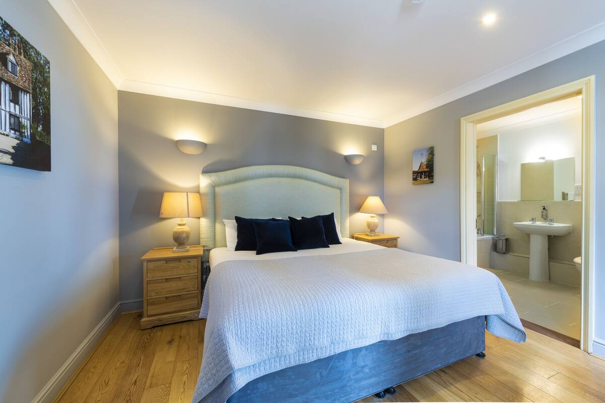 A neatly arranged bedroom with a large bed, two bedside tables with lamps, a wooden dresser, and a bathroom visible through an open door.