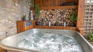Un jacuzzi con un banco de madera y plantas en macetas, frente a una pared de piedra y estantes de madera con leña.