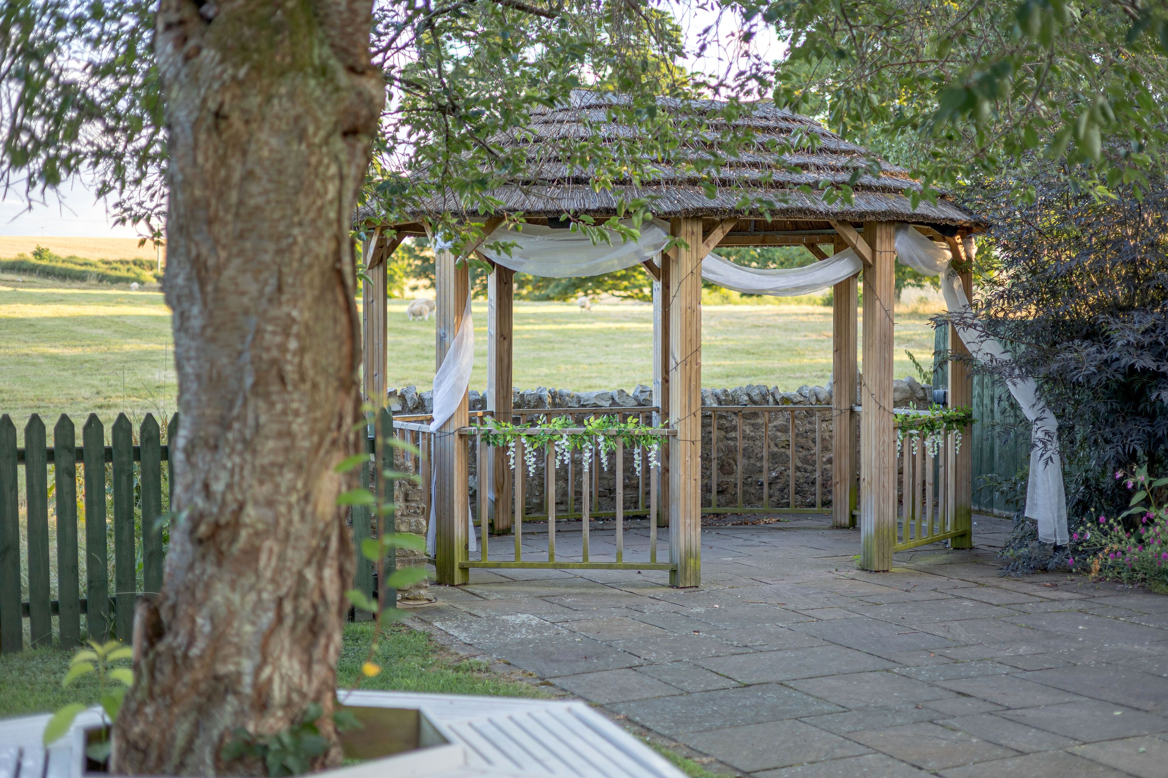 A wooden gazebo with a thatched roof, surrounded by a stone wall and a wooden fence.