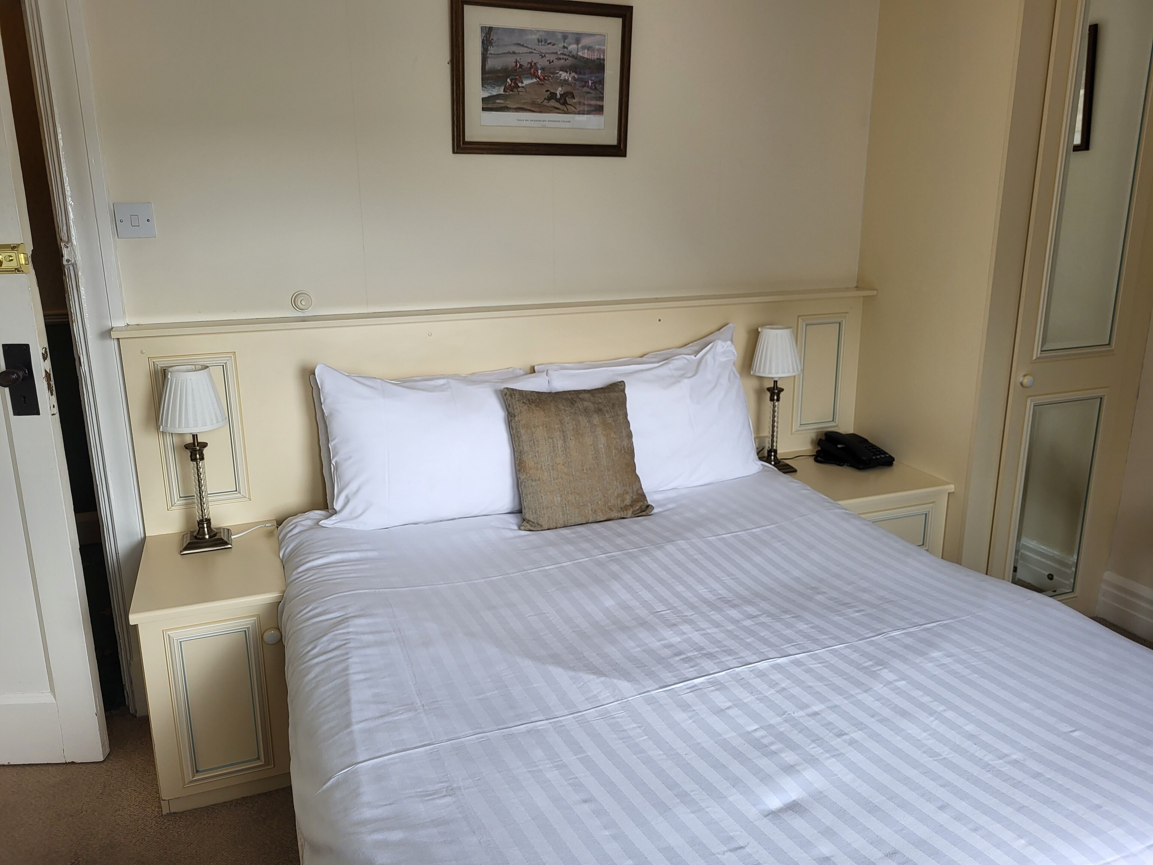 A neatly made bed with white linens and a single brown pillow, flanked by bedside tables with lamps, in a room with cream-colored walls and a framed picture above the bed.