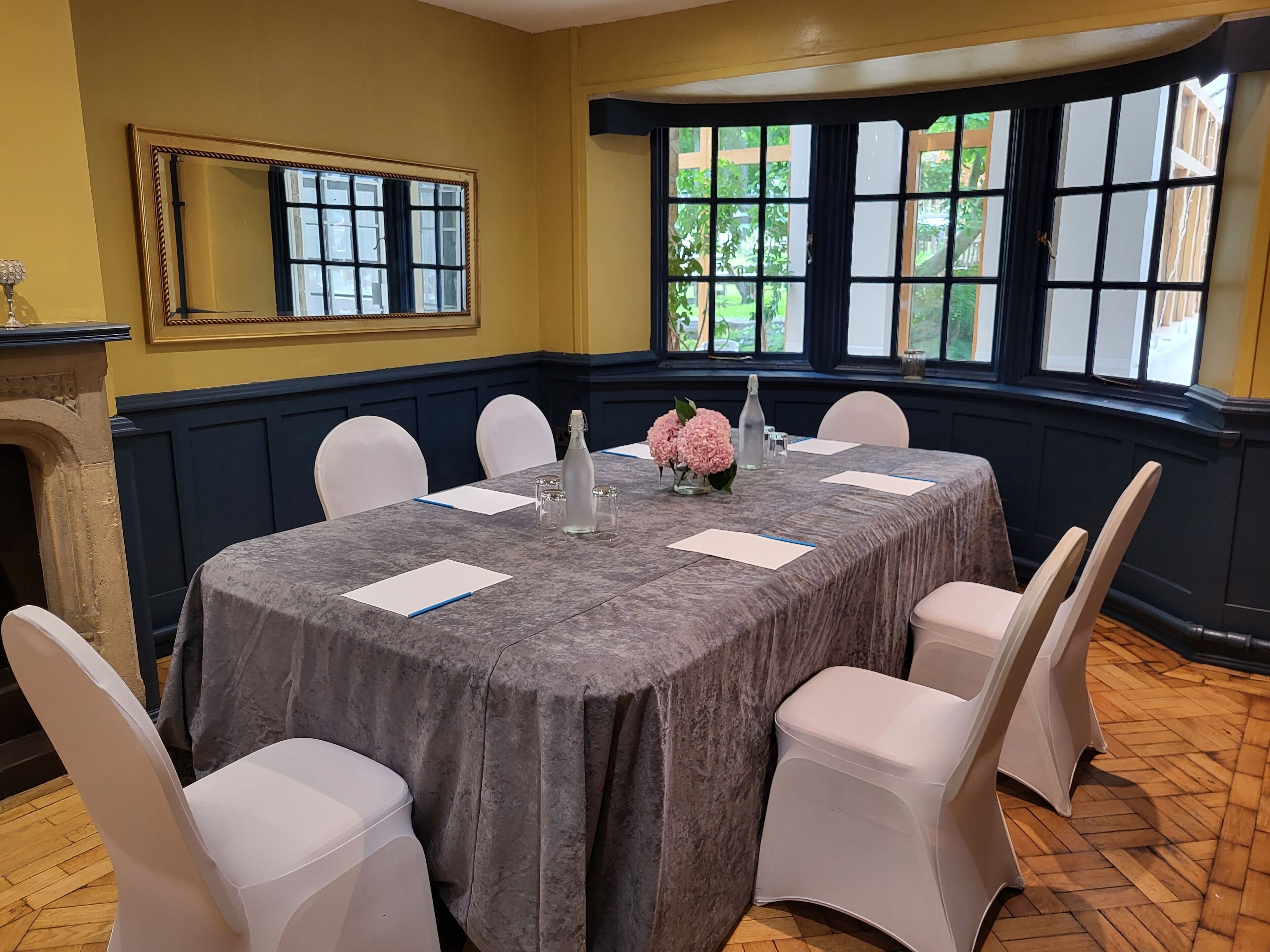 A conference room with a marble table, white chairs, a vase with pink flowers, and a view of greenery through large windows.
