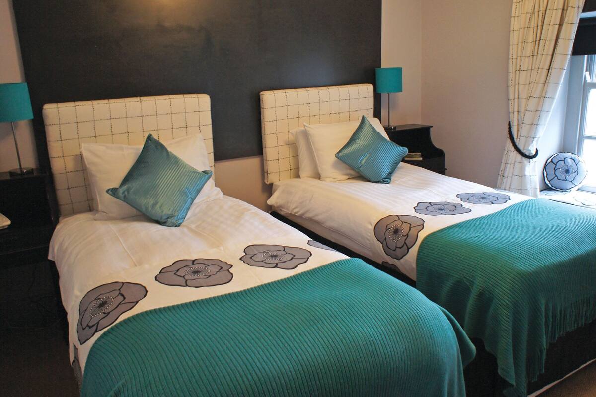 Two beds with green bedspreads and floral patterns, a phone on a nightstand, and a window with curtains.
