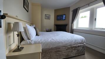Un dormitorio ordenado con cama, una mesita de noche con lámpara, chimenea, televisor y ventana con cortinas.